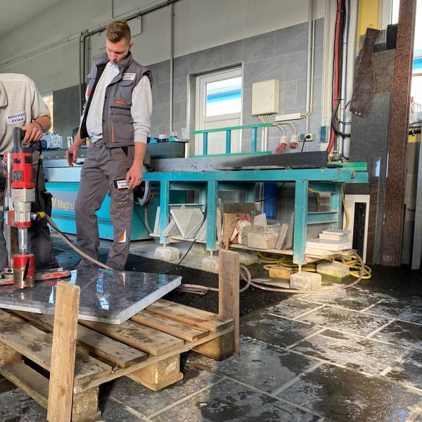 In einer Werkstatt bearbeitet der Azubi Pascal Elschenbroich eine polierte Natursteinplatte mit einem großen Bohrer. Seine Arbeit wird von einem Kollegen beobachtet.