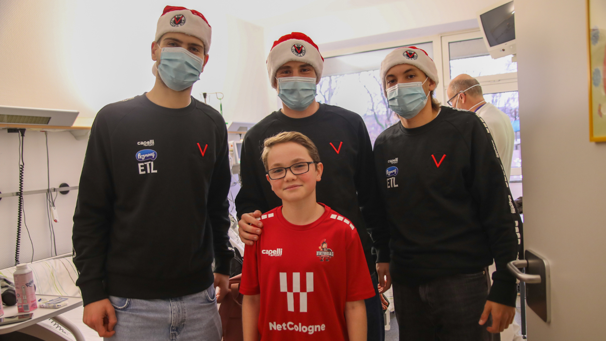 Deri junge Männer in schwarzen Pullovern, mit Weihnachtsmützen und OP-Masken und ein Junge mit Brille in einem roten T-Shirt stehen vor einem Krankenhausbett.