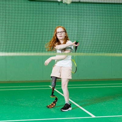 Ein Mädchen mit langen roten Haaren steht auf dem Tennisplatz. Es hat eine Beinprothese.