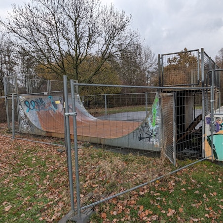 Eine mit Graffiti versehene Halfpipe für Skater hinter einem Bauzaun.