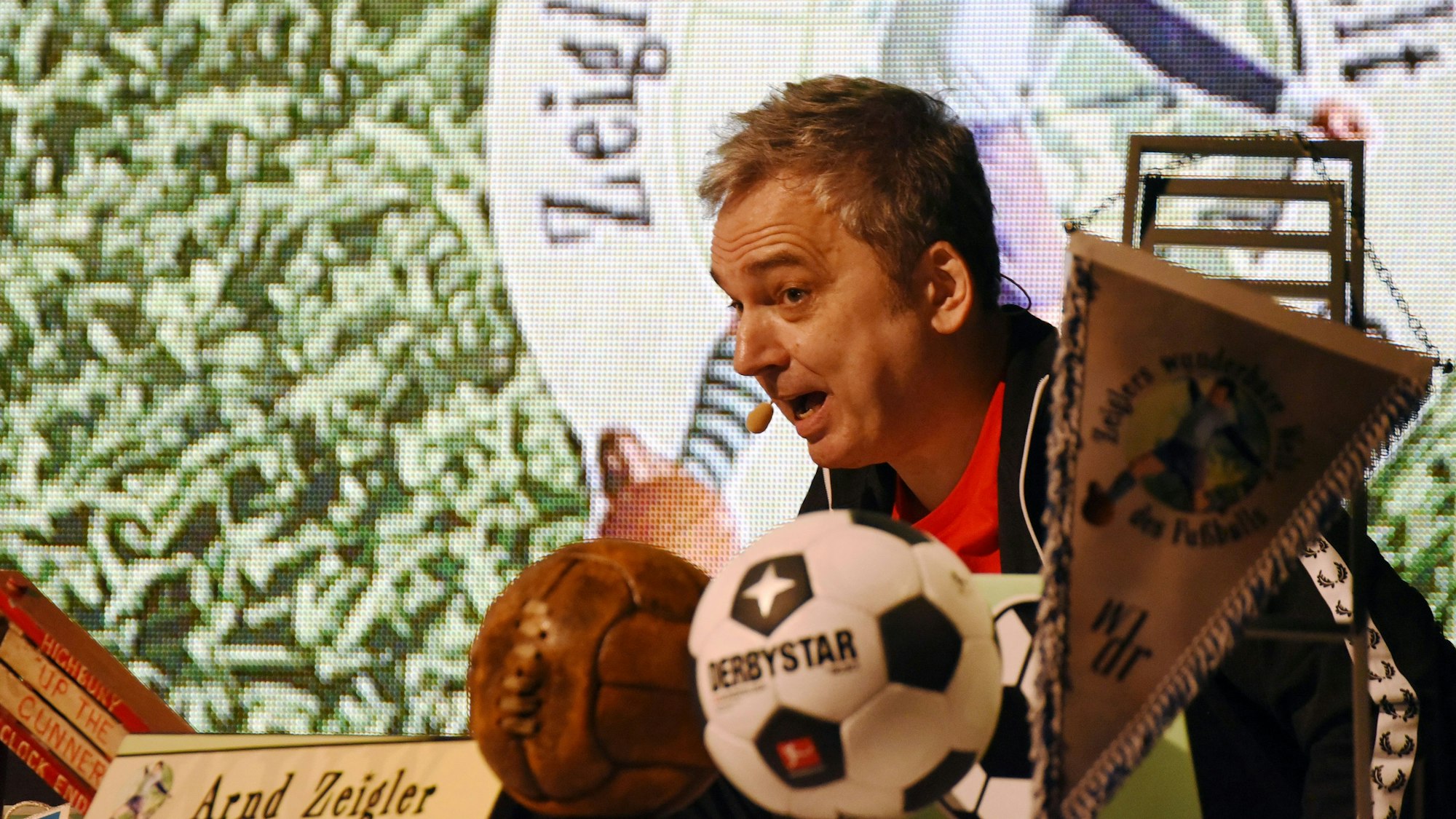 Arnd Zeigler sitzt an dem aus dem Fernsehen bekannten Schreibtisch und spricht zum Publikum.