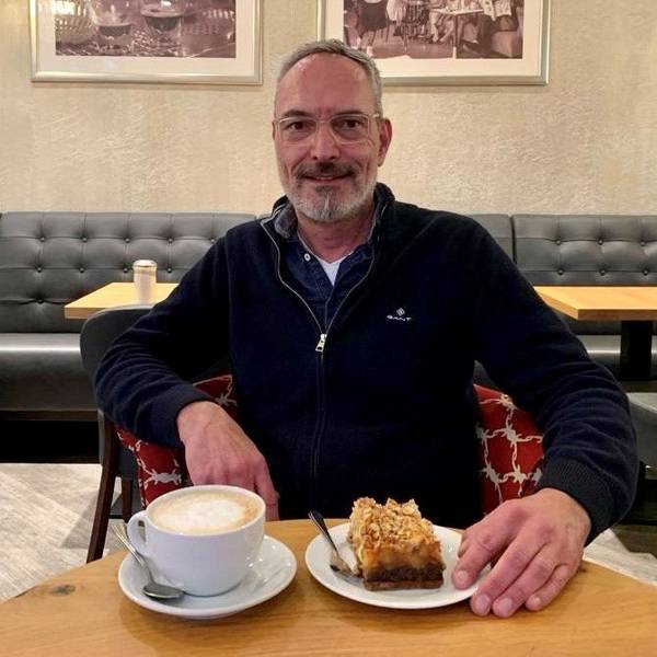 Zu sehen ist ein Mann in einem Café, der vor sich einen Kaffee und ein Stück Kuchen auf dem Tisch stehen hat.