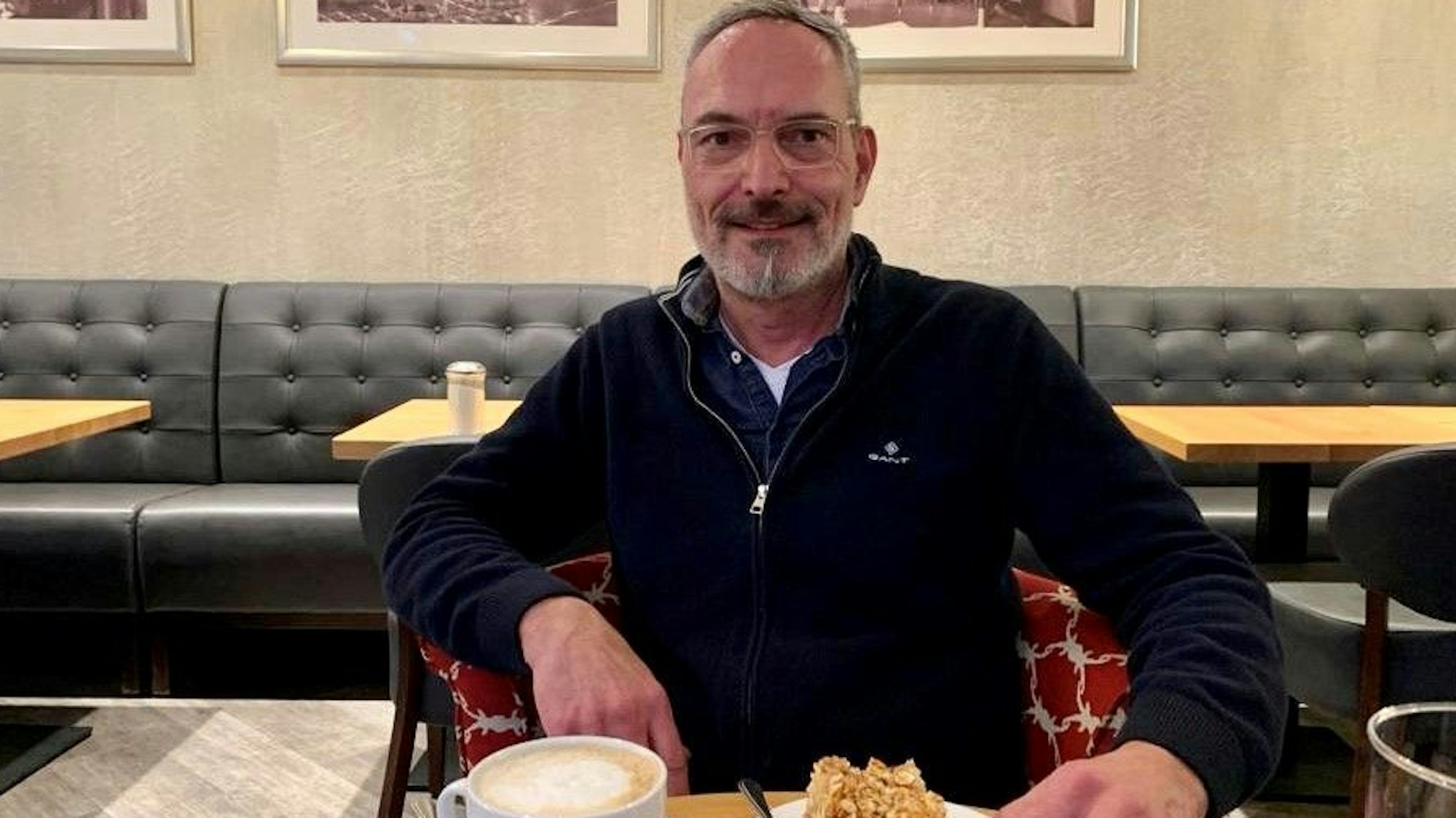 Zu sehen ist ein Mann in einem Café, der vor sich einen Kaffee und ein Stück Kuchen auf dem Tisch stehen hat.