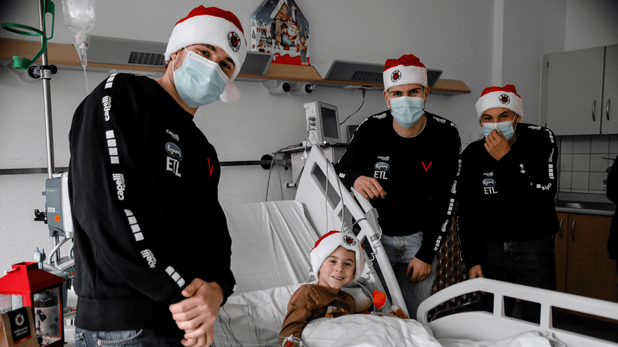 Drei junge Männer in schwarzen Pullis und mit Weihnachtsmützen stehen am Bett eines Kindes mit Weihnachtsmütze.