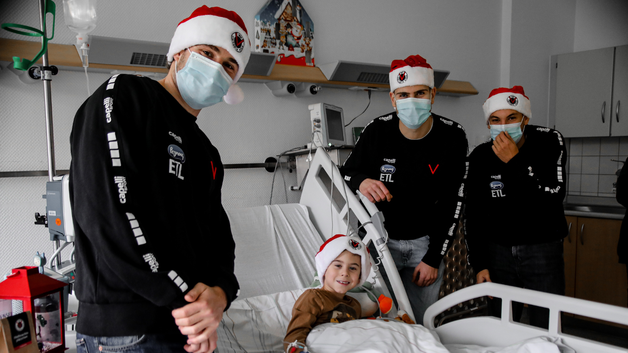 Drei junge Männer in schwarzen Pullis und mit Weihnachtsmützen stehen am Bett eines Kindes mit Weihnachtsmütze.