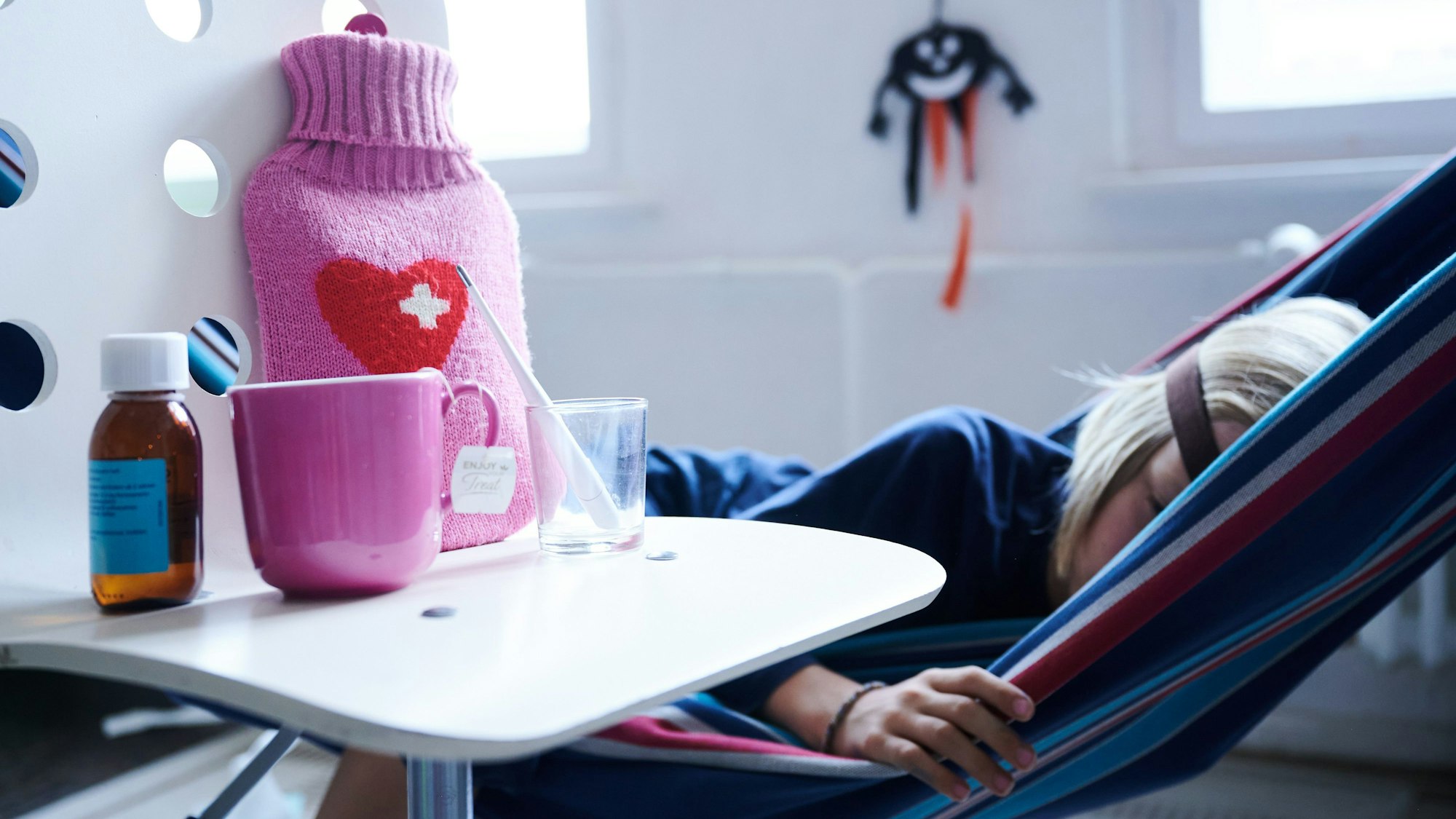 ARCHIV - 11.09.2021, Berlin: ILLUSTRATION - Fieberthermometer, Hustensaft, Wärmflasche und eine Tasse Tee stehen auf einem Stuhl, während ein Kind im Hintergrund in einer Hängematte liegt. In Deutschland hat nach Einschätzung des Robert Koch-Instituts (RKI) die Welle der RSV-bedingten Atemwegserkrankungen begonnen. (zu dpa «Atemwegserkrankungen: RSV-Welle in Deutschland hat laut RKI begonnen») Foto: Annette Riedl/dpa +++ dpa-Bildfunk +++