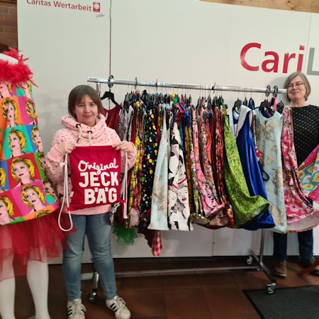 Eine junge Frau hält eine Stofftasche mit dem Schriftzug Original Jeck Bäg, eine ältere Frau steht neben einer Kleiderstange mit vielen Kleidern in verschiedenen Farben.