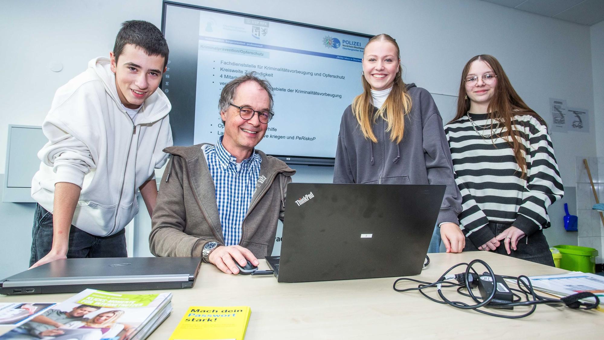 An einem Tisch sitzt ein Mann vor einem Notebook, links und rechts neben ihm stehen zwei Schülerinnen und ein Schüler.