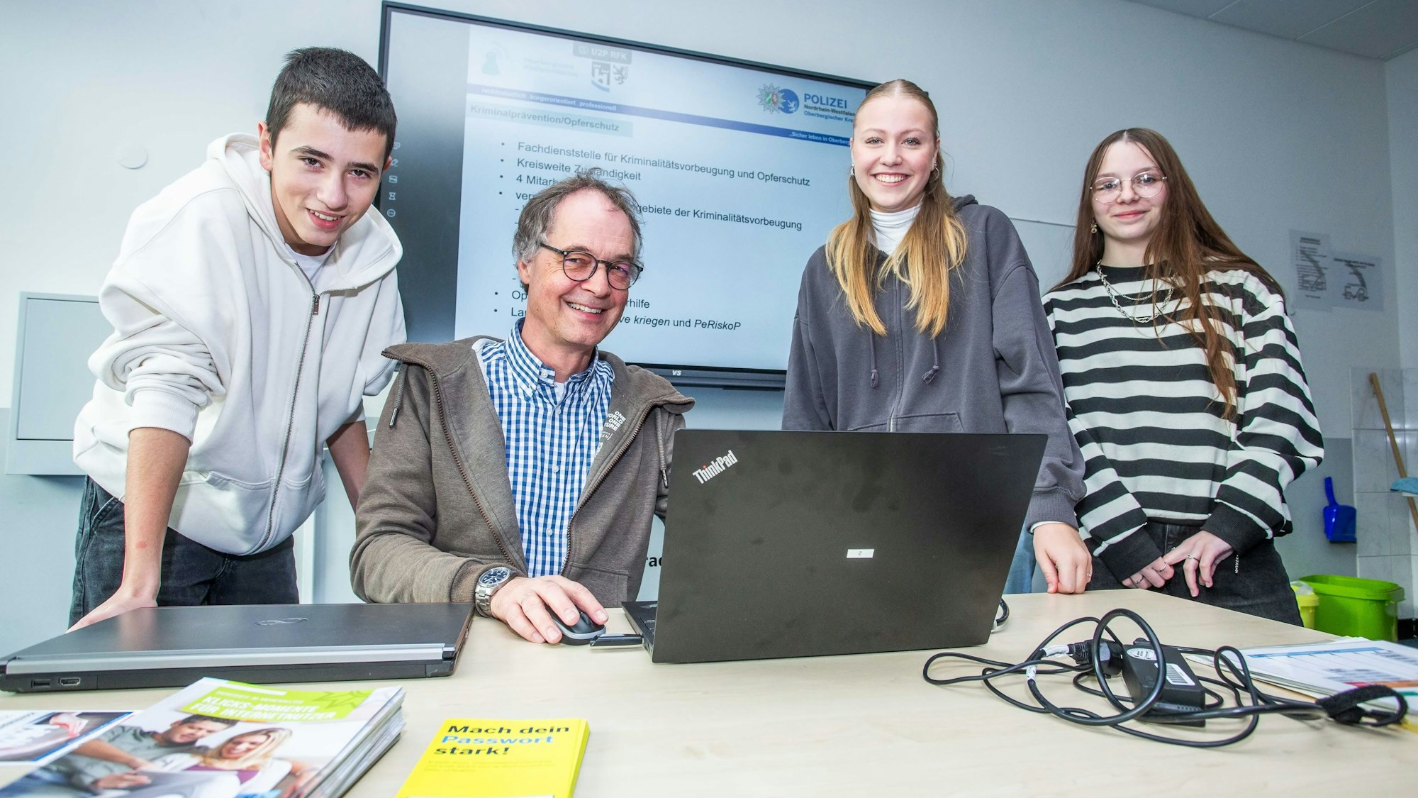 An einem Tisch sitzt ein Mann vor einem Notebook, links und rechts neben ihm stehen zwei Schülerinnen und ein Schüler.