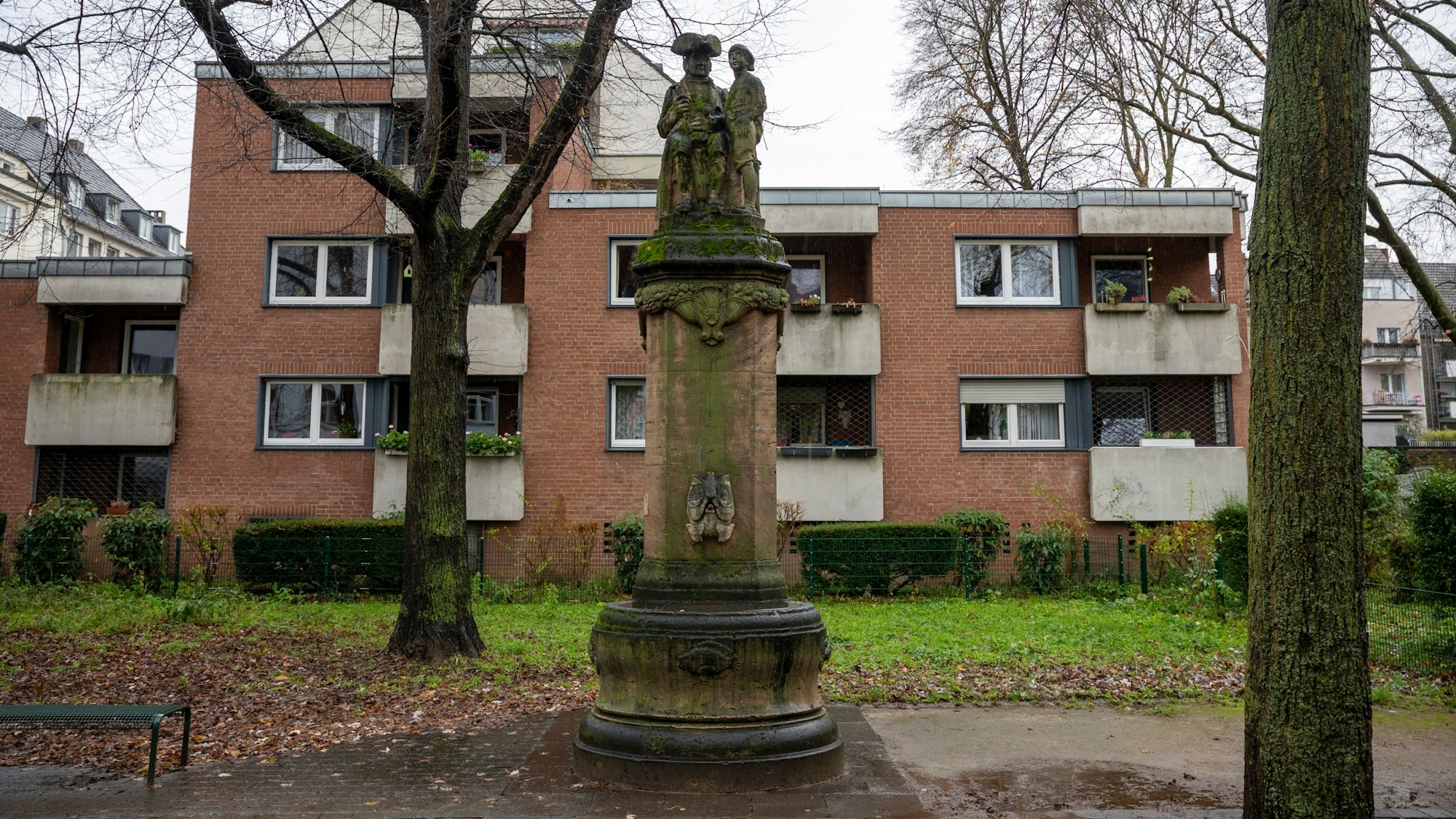 Zu sehen ist eine Brunnensäule mit zwei Skulpturen darauf, dahinter ein Wohnhaus.