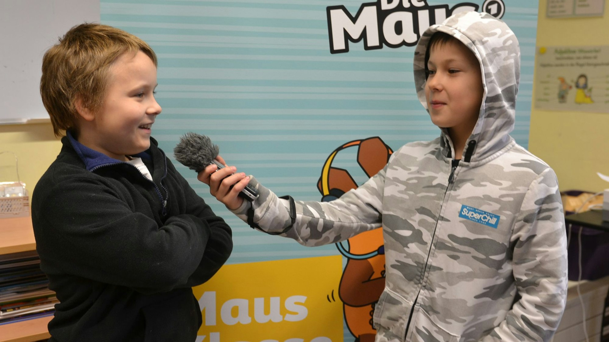 Zwei Schüler stehen vor einem Plakat der Sendung mit der Maus. Einer hält dem anderen ein Mikrofon hin.