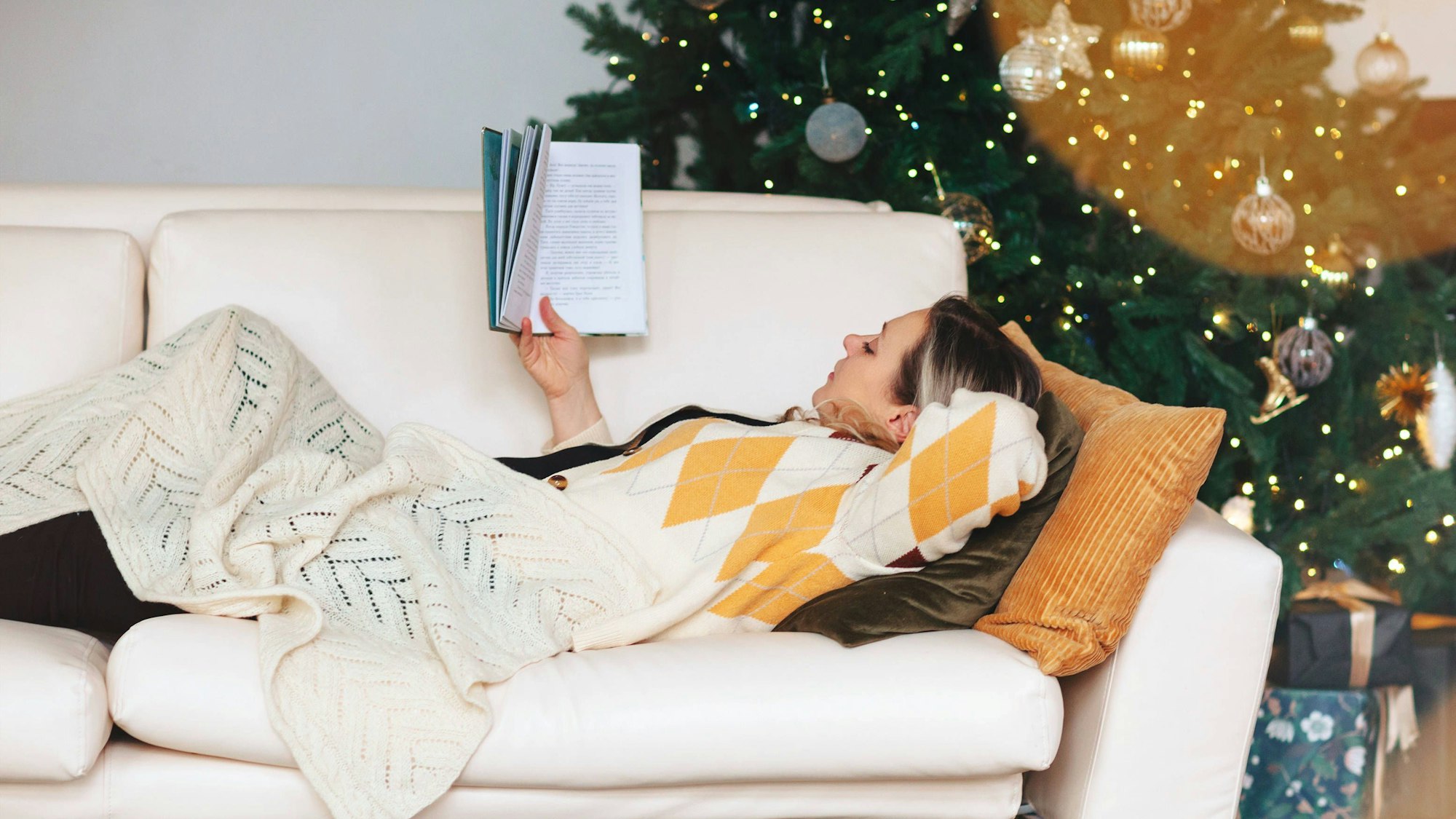 Eine Frau liest, während sie auf dem Sofa im Wohnzimmer mit geschmücktem Weihnachtsbaum liegt