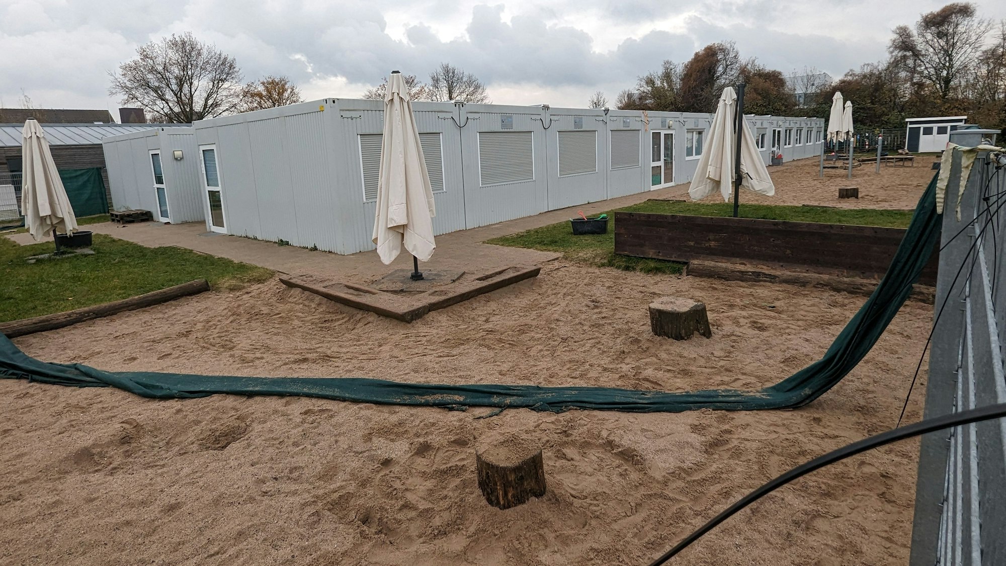 Provisorisch hergerichtete Sandkästen mit Baumstümpfen vor einer Containeranlage.