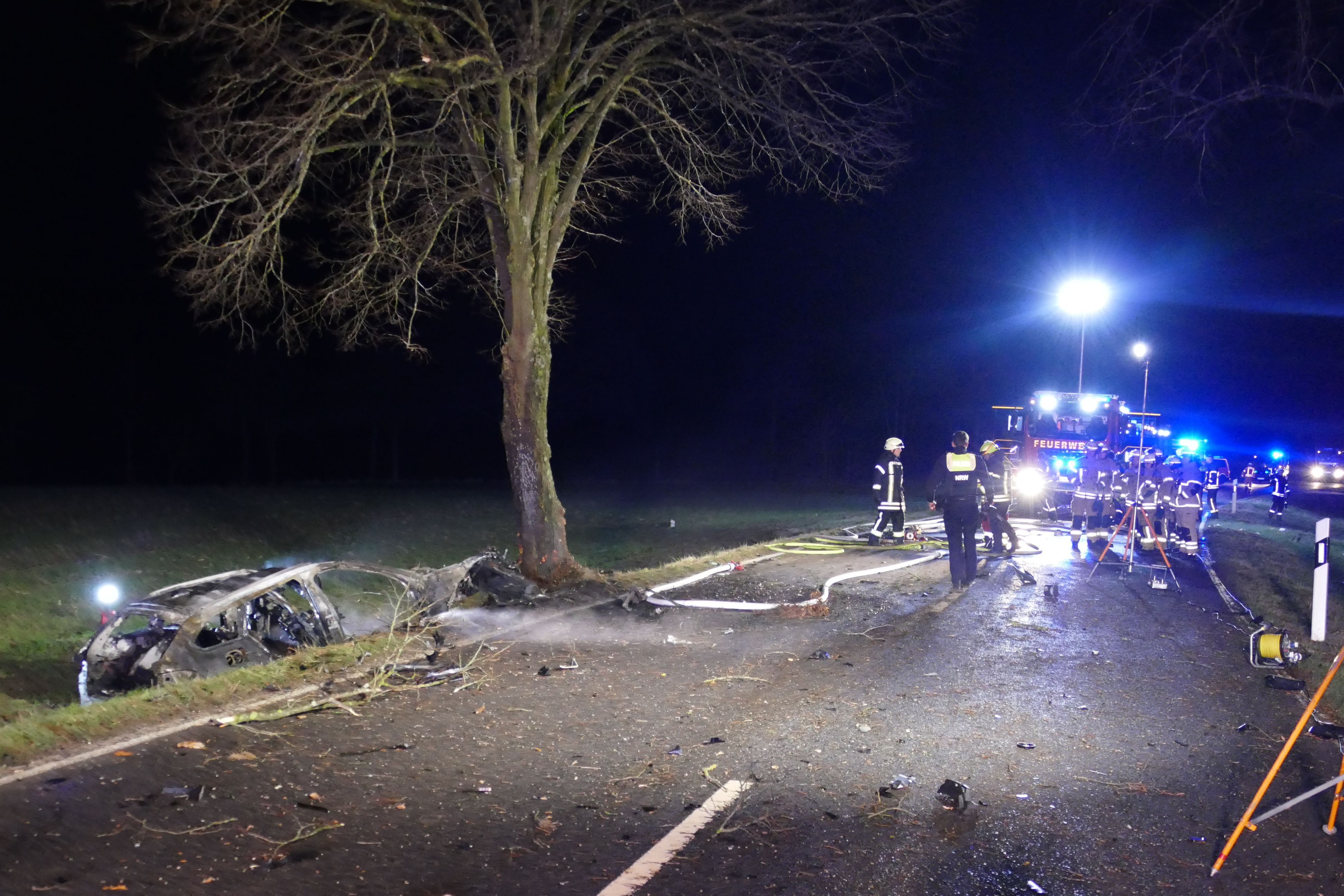 Trümmer liegen auf der Fahrbahn verteilt, das ausgebrannte Wrack liegt neben dem Baum.