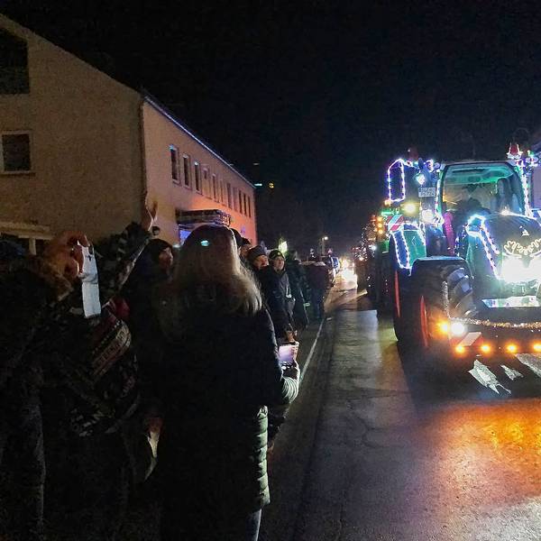Ein mit Lichterketten behangener Traktor fährt über eine Straße, links und rechts stehen Schaulustige.