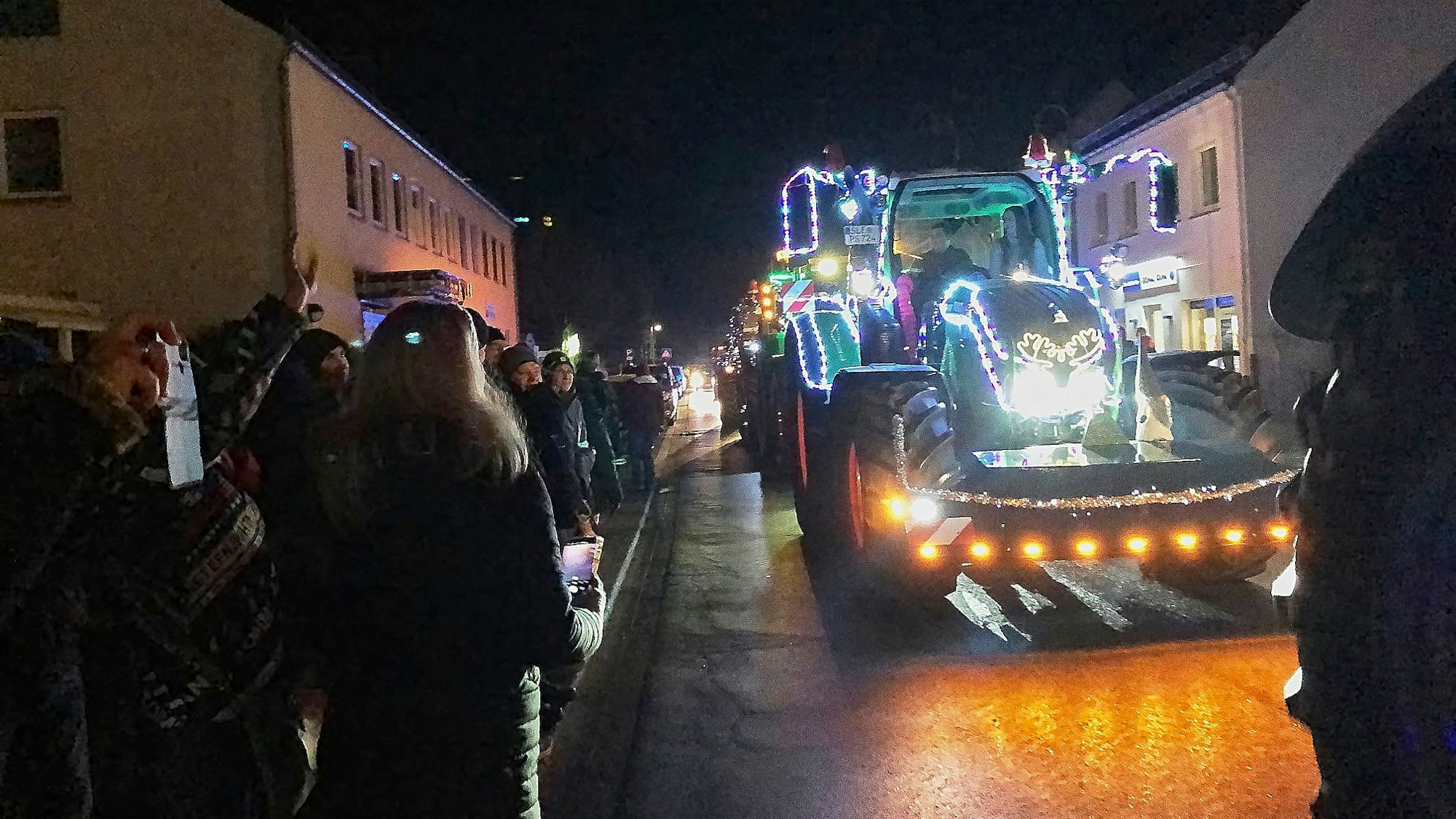 Ein mit Lichterketten behangener Traktor fährt über eine Straße, links und rechts stehen Schaulustige.