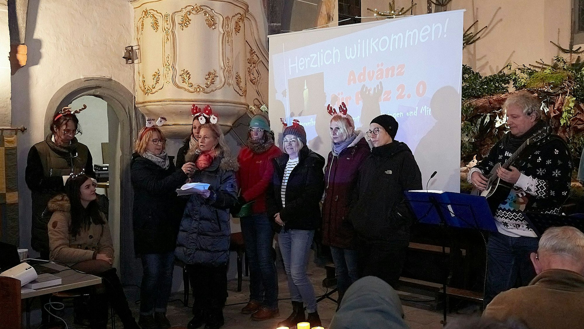 Eine Gruppe Frauen mit weihnachtlichen Haarreifen steht im Altarraum vor einer Leinwand. Im Hintergrund sind Weihnachtsbäume zu erkennen.