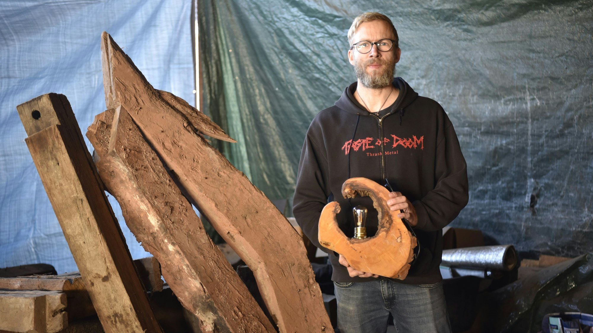 Björn Schütz steht in einem Schuppen neben alten Holzbalken. In der Hand hält er eine Lampe aus einer Baumscheibe.