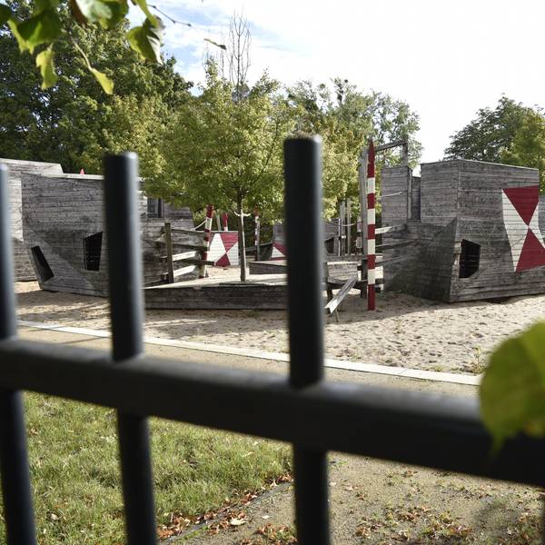 Hinter einem Zaun sind Spielgeräte aus Holz zu erkennen.