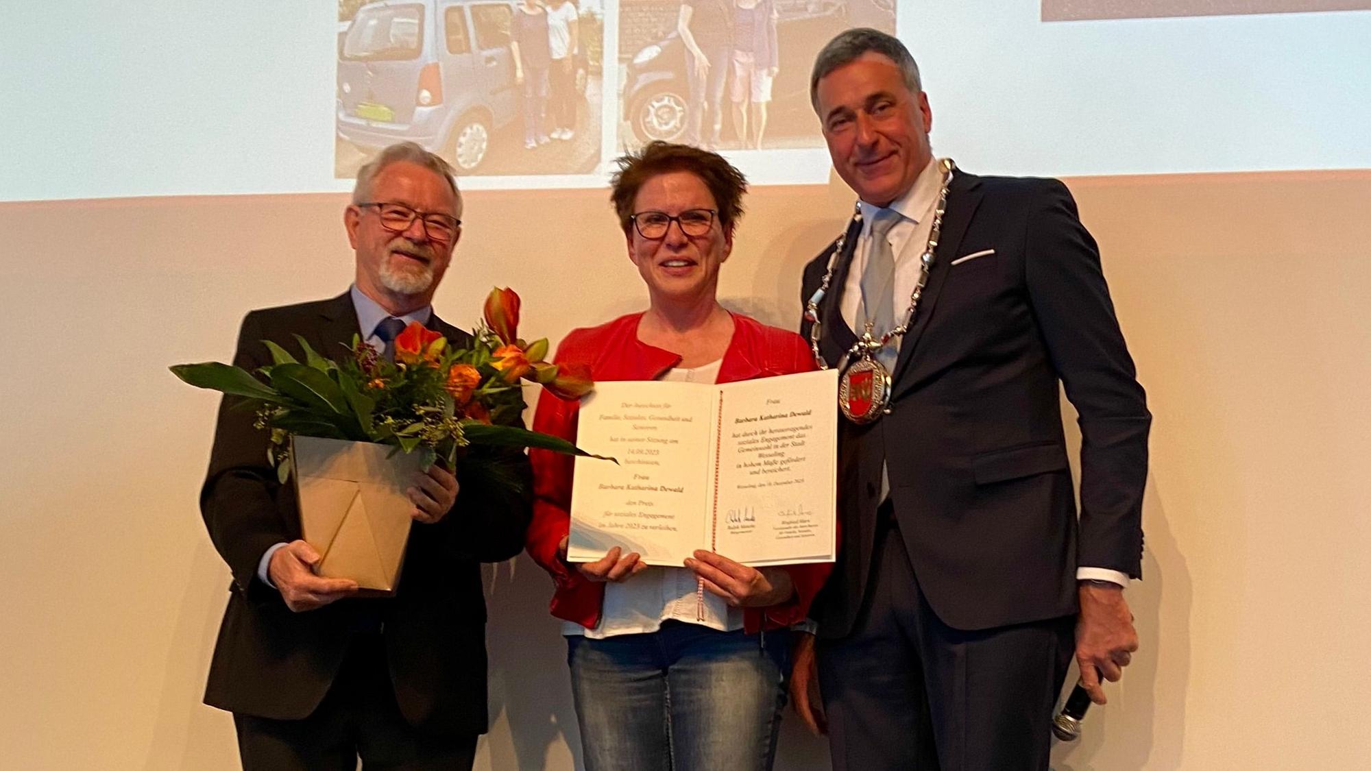 Zwei Männer überreichen einer Frau eine Urkunde und Blumen.
