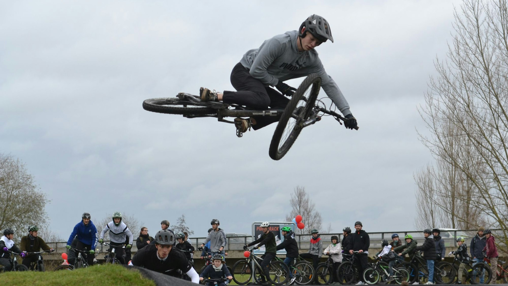Ein Jugendlicher im grauen Sweatshirt führt einen Fahrrad-Trick in der Luft vor.