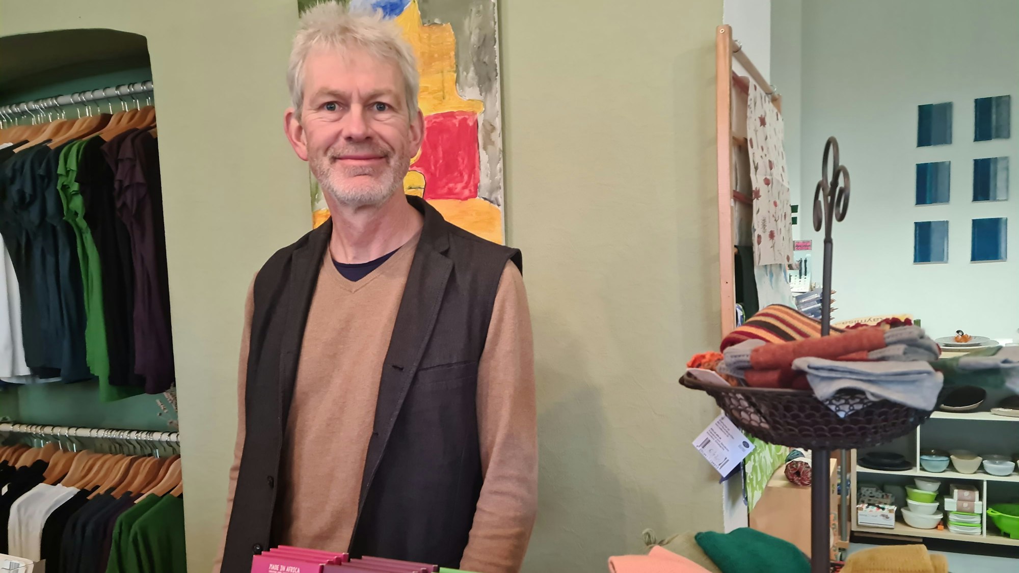 Ein Mann mit grauem Haar und Bart steht in einem Laden, in dem T-Shirts auf Bügeln hängen und Pulswärmer auf einer Etagere liegen.