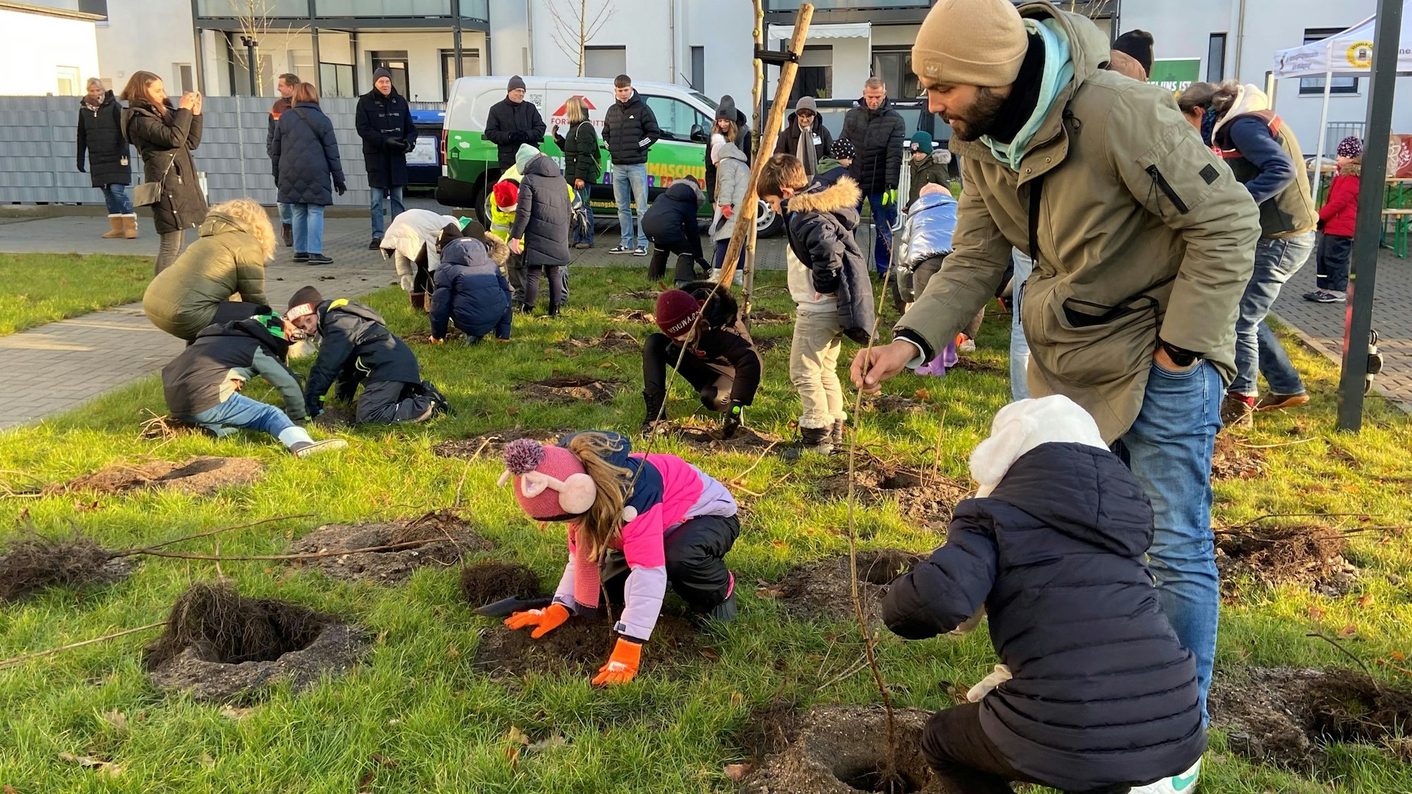 Mehrere Kinder hocken auf einer Rasenfläche vor Erdlöchern und pflanzen Sträucher ein.
