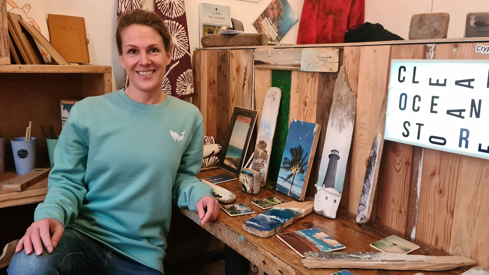 Susanne Weichold, eine Frau mit hochgebundenen Haaren und türkisem Pullover, sitzt vor bedrucktem Treibholz in einem Ladenlokal.