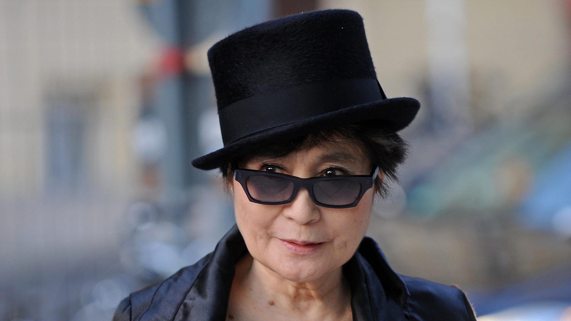 Yoko Ono steht vor einem verschwommenem Hintergrund und trägt einen schwarzen Zylinder, schwarze Sonnenbrille und Lederjacke. Sie schaut über die Ränder ihrer Brille und lächelt.