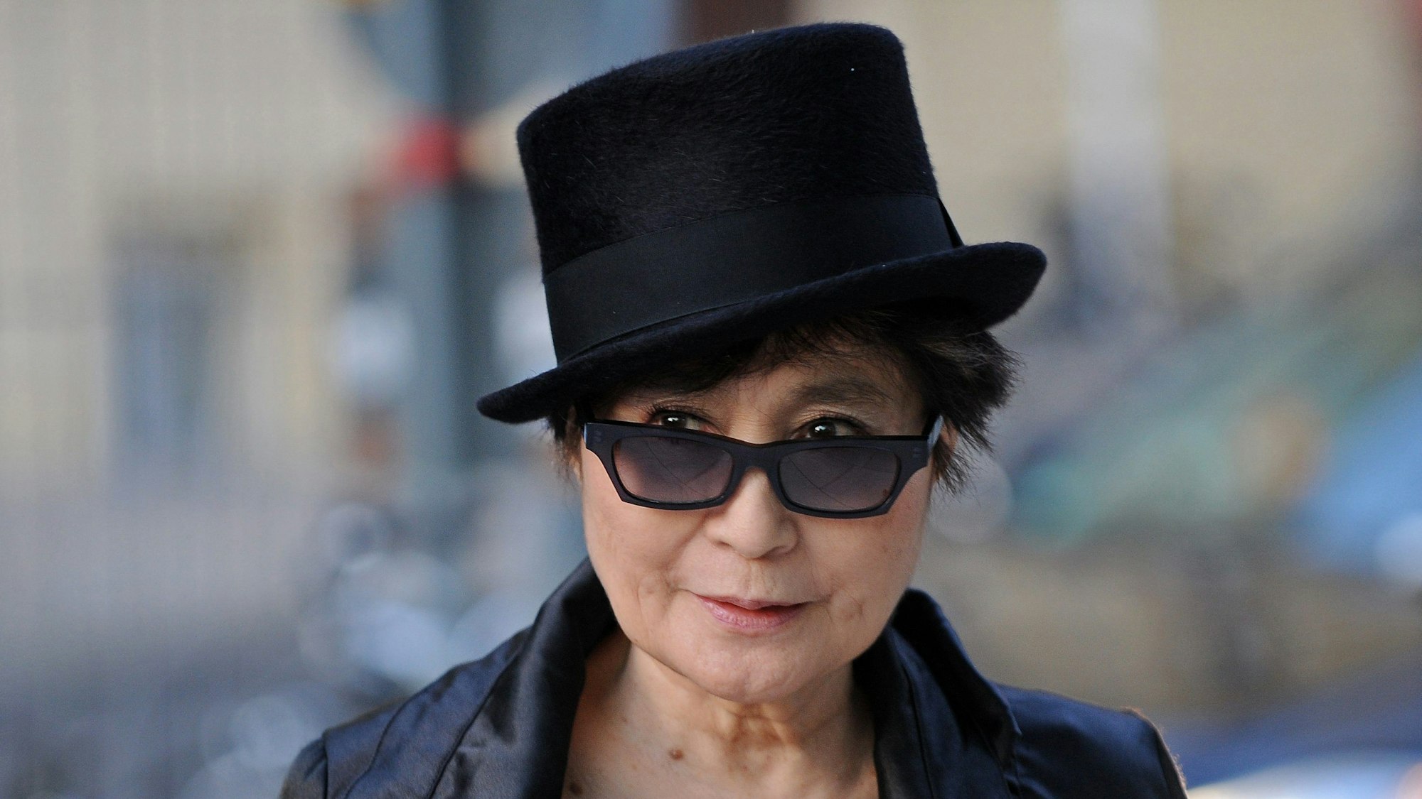 Yoko Ono steht vor einem verschwommenem Hintergrund und trägt einen schwarzen Zylinder, schwarze Sonnenbrille und Lederjacke. Sie schaut über die Ränder ihrer Brille und lächelt.