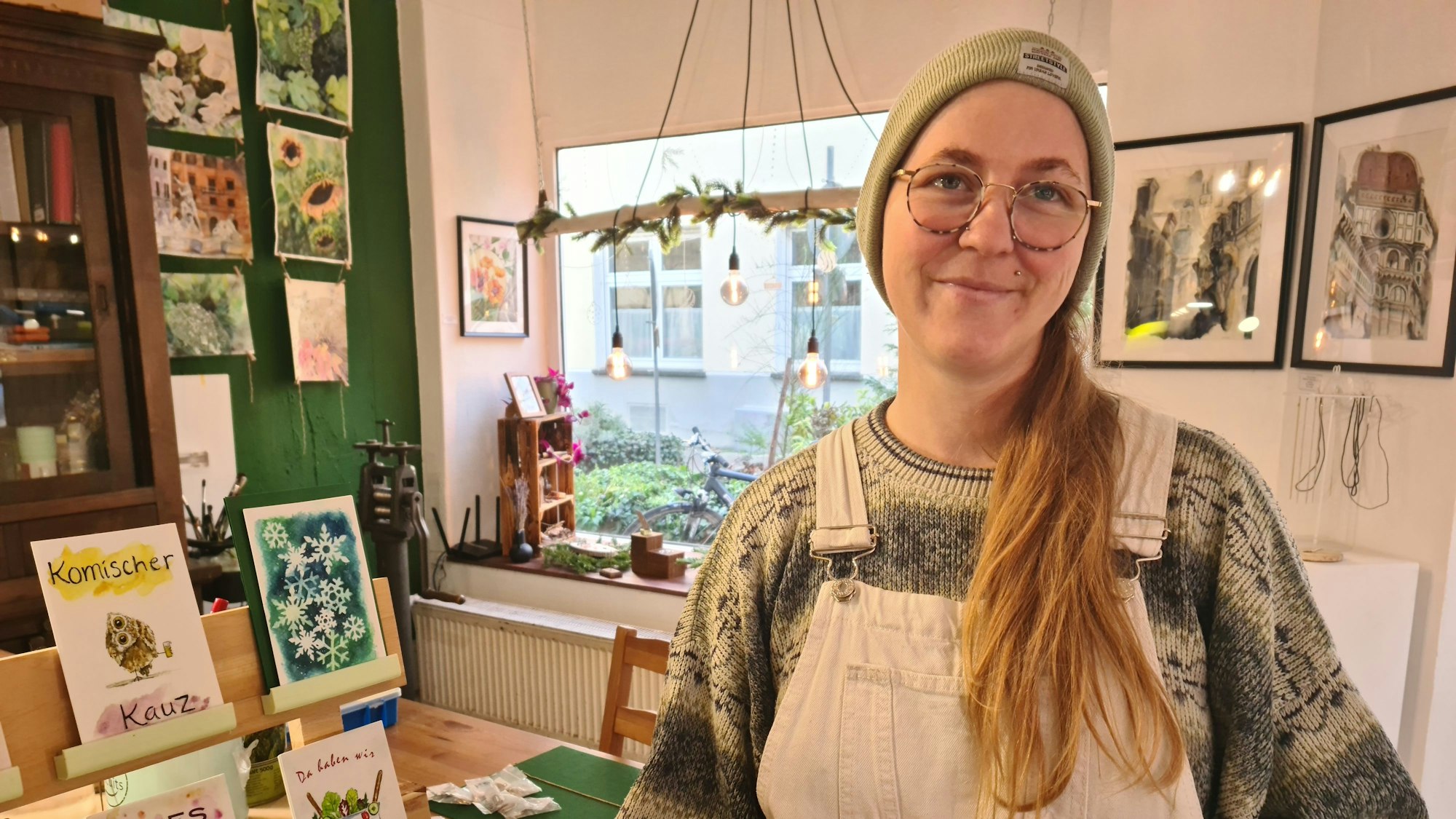 Eine Frau mit langem Zopf, Mütze und Brille steht in einem Raum, der mit Bildern dekoriert ist. Sie hält einen Schaukasten mit Ohrringen und Halsketten.