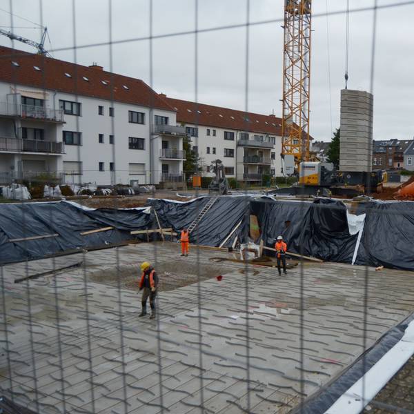 Drei Arbeiter stehen in einer Baugrube, die für die Errichtung eines Mehrfamilienhauses ausgehoben worden ist. Im Hintergrund weitere Wohngebäude und ein Baukran.