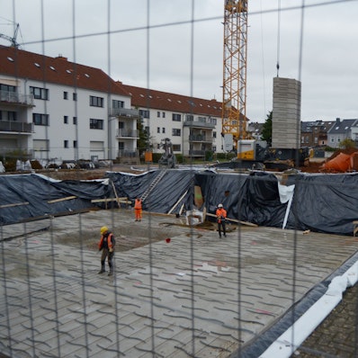 Drei Arbeiter stehen in einer Baugrube, die für die Errichtung eines Mehrfamilienhauses ausgehoben worden ist. Im Hintergrund weitere Wohngebäude und ein Baukran.