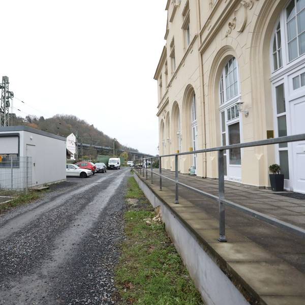 Ein geschotterter Weg führt zwischen dem Bahnübergang Drachenfelsstraße und einem Gebäude entlang.
