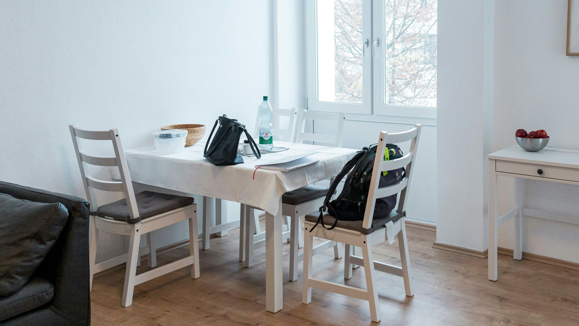 Zu sehen ist ein helles, schlichtes Wohnzimmer mit weißen Möbeln, Holzboden und einer schwarzen Ledercouch.