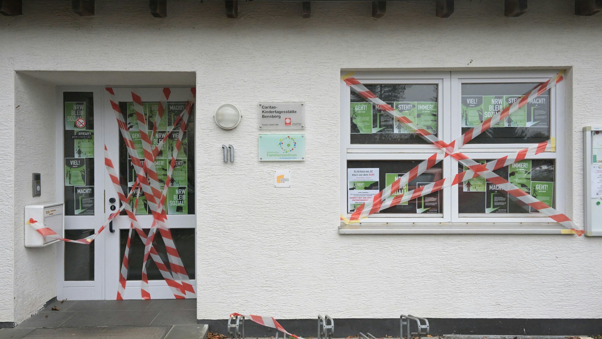 Die Eingangstür und ein Fenster des weißen Kita-Gebäudes sind mit rot-weißem Flatterband beklebt. Im Fenster hängen DinA 4 große Protestplakate.