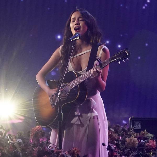 Olivia Rodrigo steht mit einer Gitarre auf der Bühne, auf welcher Blumen stehen und singt. Sie steht im Scheinwerferlicht vor einem blauem Hintergrund.