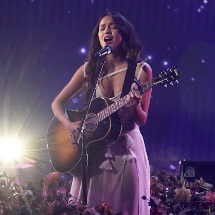 Olivia Rodrigo steht mit einer Gitarre auf der Bühne, auf welcher Blumen stehen und singt. Sie steht im Scheinwerferlicht vor einem blauem Hintergrund.