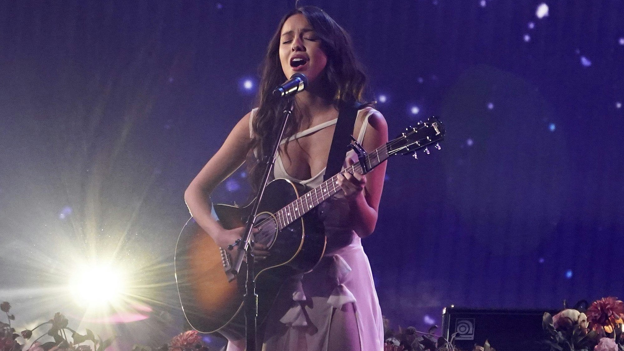 Olivia Rodrigo steht mit einer Gitarre auf der Bühne, auf welcher Blumen stehen und singt. Sie steht im Scheinwerferlicht vor einem blauem Hintergrund.