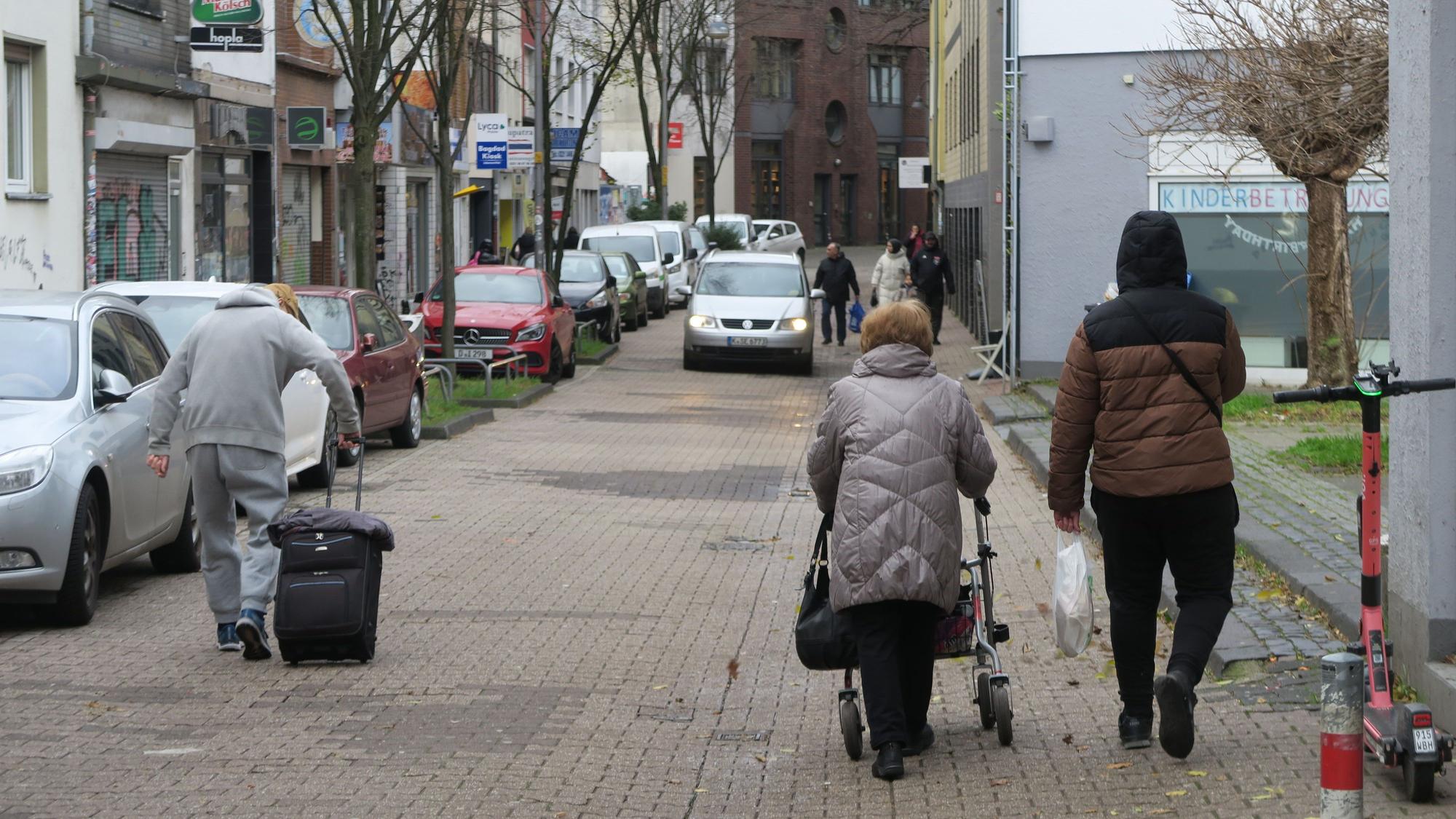 Drei Fußgänger gehen auf der Steprathstraße, eine Frau schiebt einen Rollator, ein Mann zieht einen Koffer hinter sich her. Ihnen kommt ein Auto entgegen.