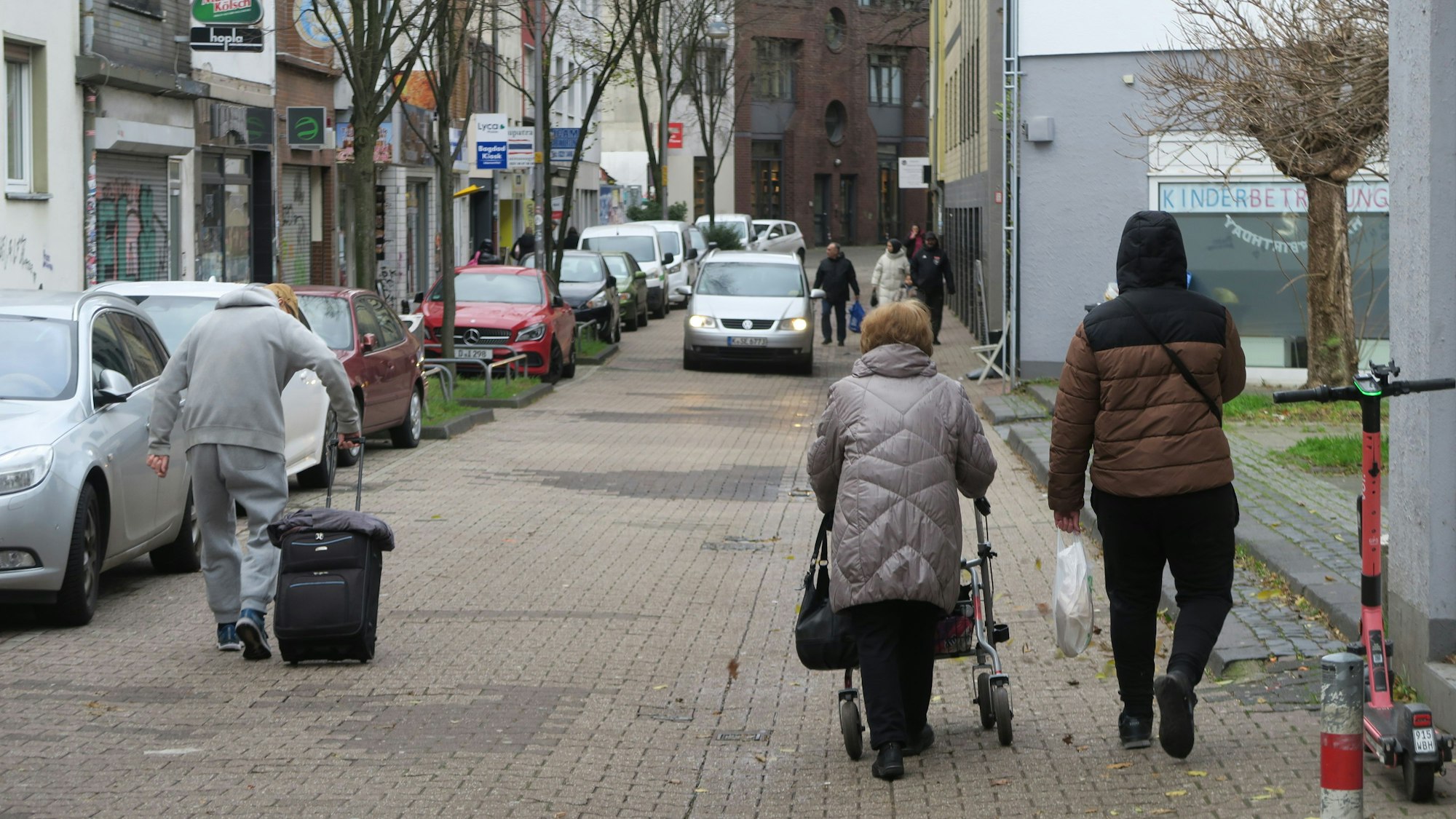 Drei Fußgänger gehen auf der Steprathstraße, eine Frau schiebt einen Rollator, ein Mann zieht einen Koffer hinter sich her. Ihnen kommt ein Auto entgegen.