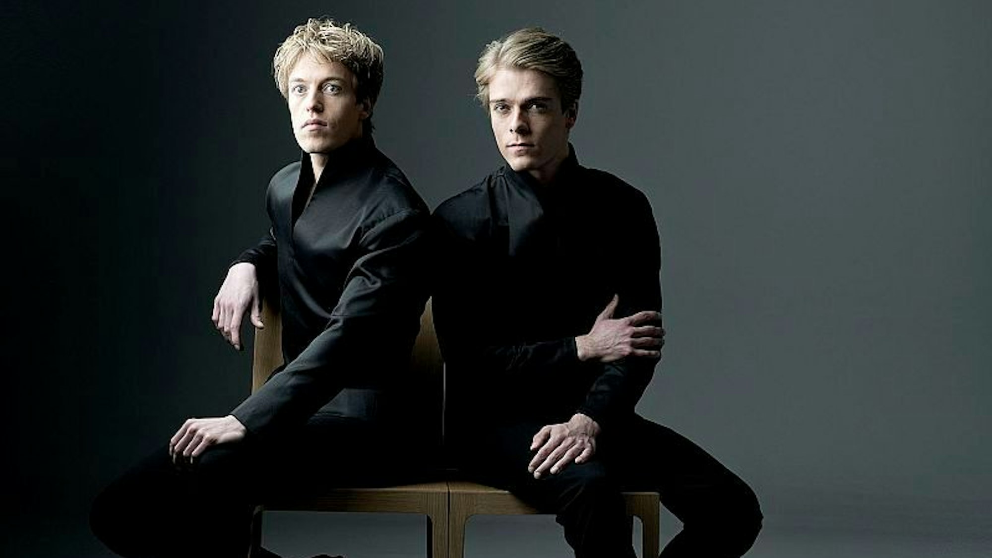 Lucas und Arthur Jussen Pianisten. Sie tragen schwarze Anzüge und sitzen auf zwei Stühlen nebeneinander. Der Hintergrund ist grau.