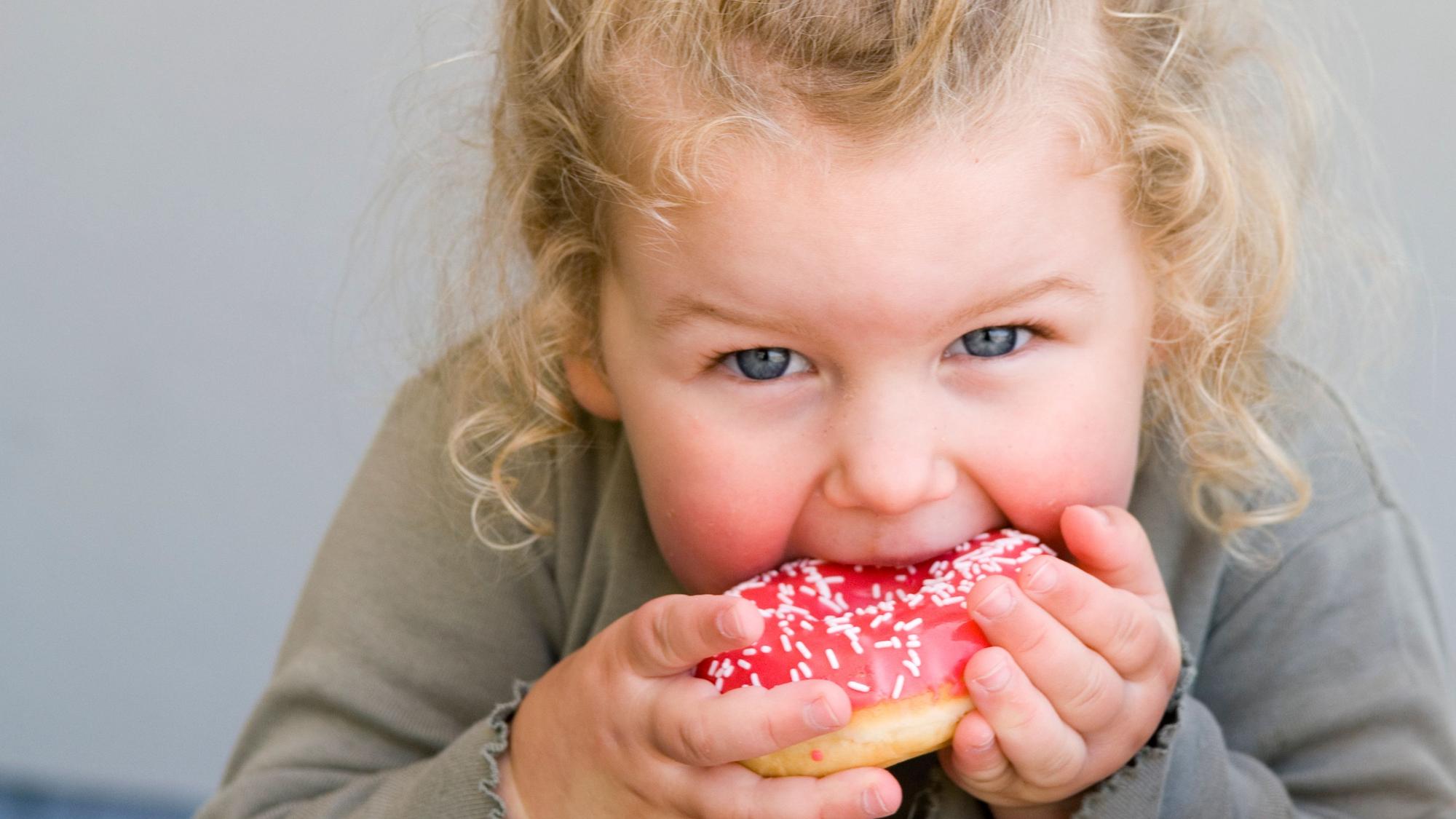 Ein kleines Mädchen isst einen pinken Donut. Symbolbild
