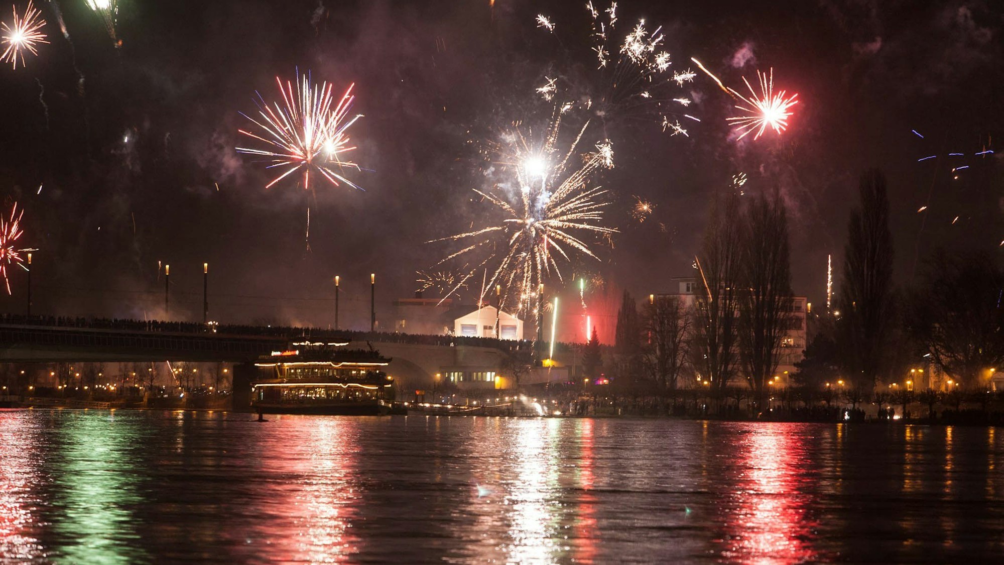 Silvesterfeuerwerk vom Bonner Rheinufer: Feuerwerk erleuchtet den dunklen Nachthimmel über dem Rhein und der Kennedybrücke