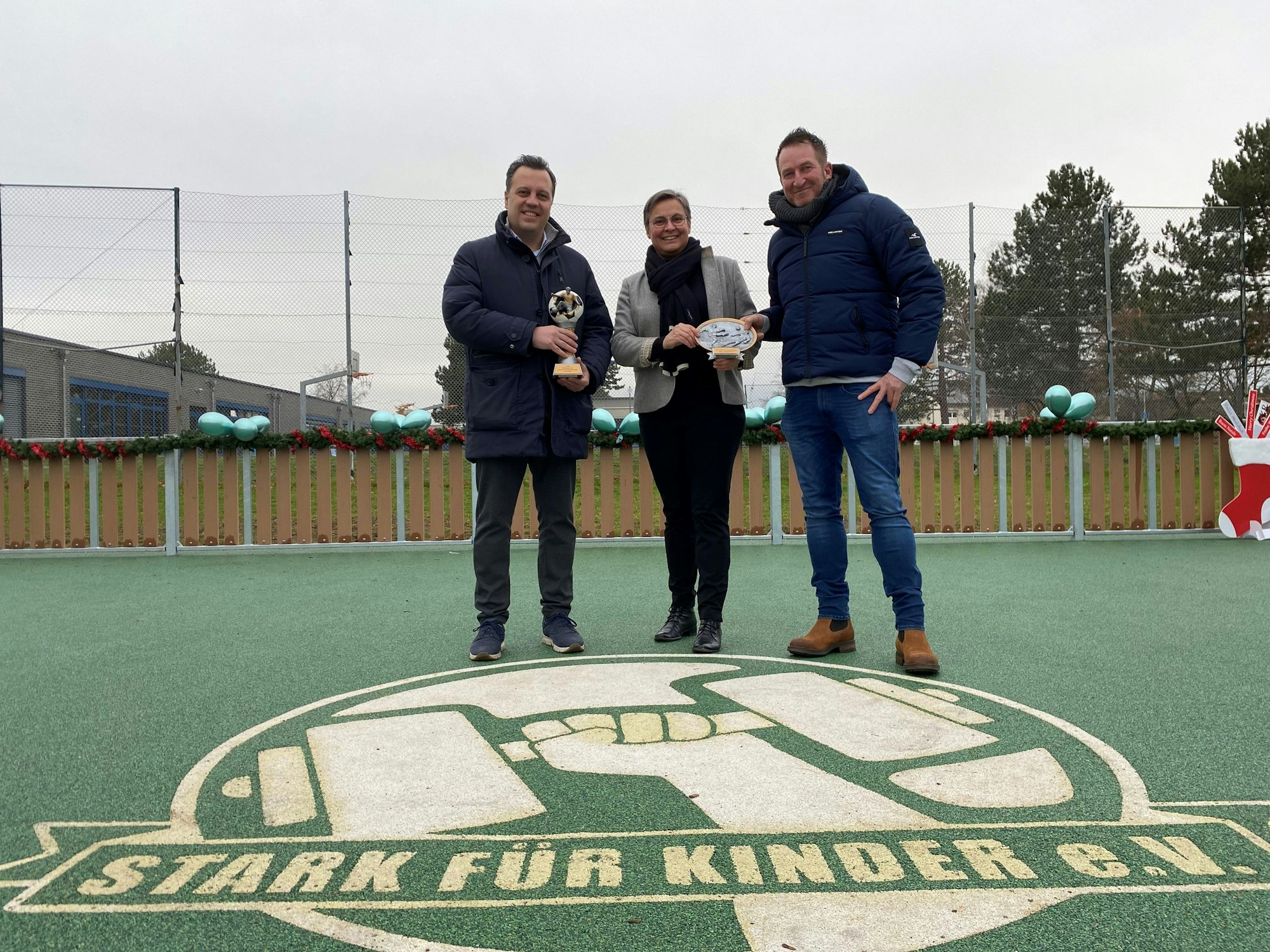 Bürgermeister Sacha Reichelt, Schulleiterin Karin Bossant und der Vereinsvorsitzende Volker Zimmer stehen hinter einem auf den grünen Tartan-Platz gedruckten Logo. Auf dem Logo ist eine Hand zu sehen, die eine Hantel hält. Die Aufschrift: „Stark für Kinder“.