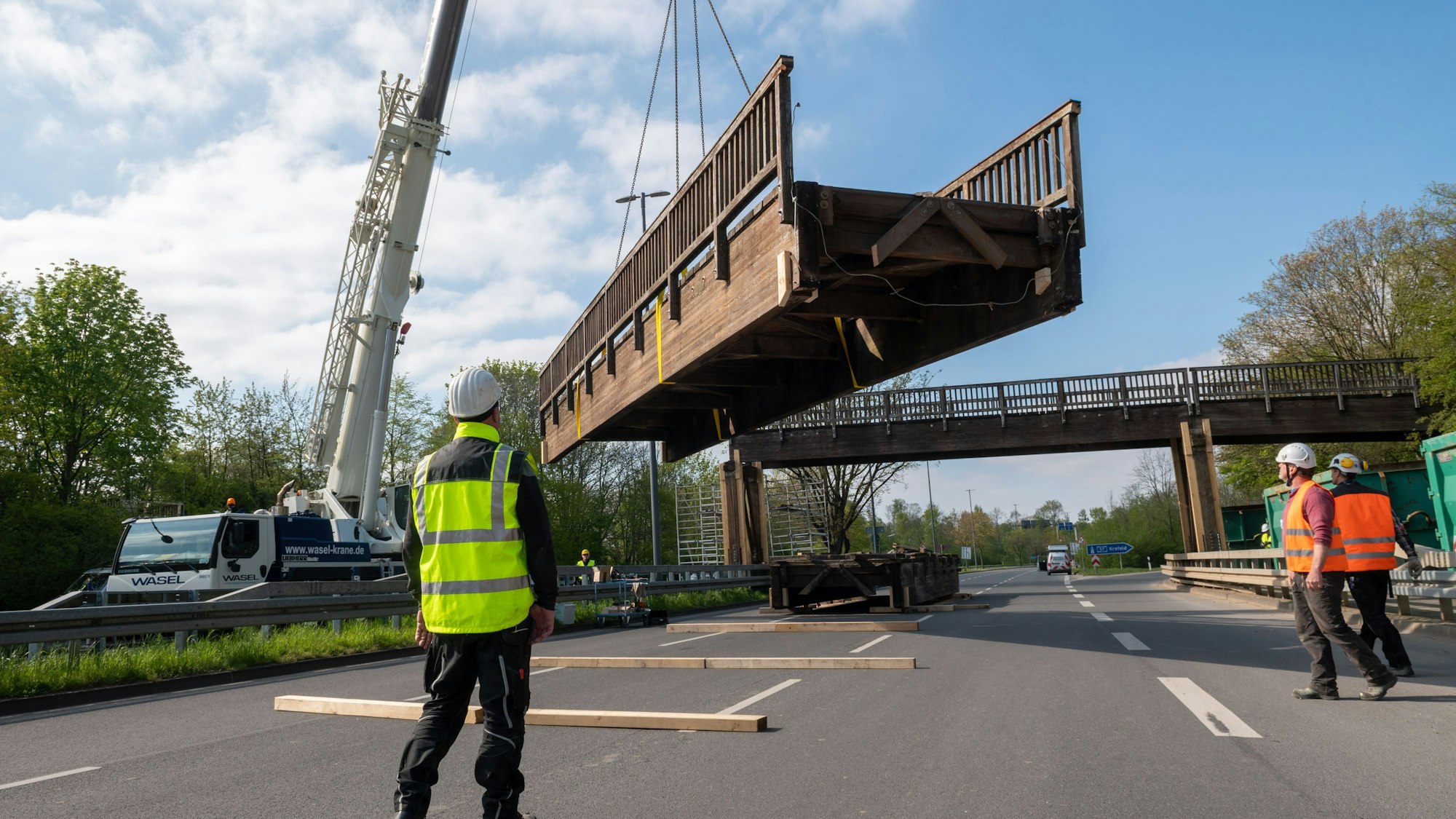 Ein Brückenteil wird von einem Kran angehoben, Arbeiter stehen auf einer mehrspurigen Straße.