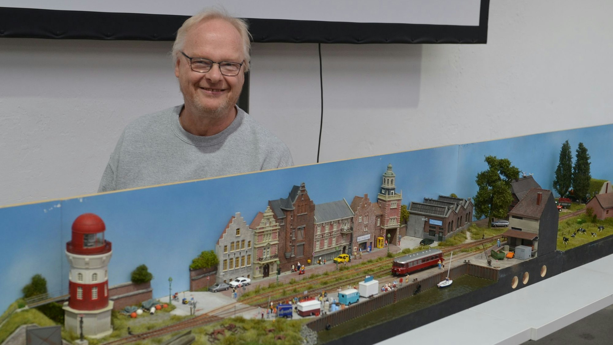 Der Modellbauer Wilfried Kurth steht vor seiner Anlage mit Bahngleisen, Häusern, einem Hafen und einem Leuchtturm.