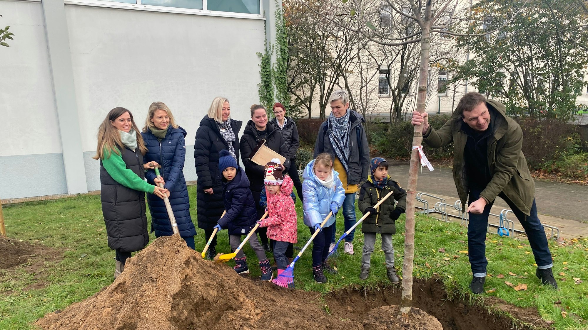 Sieben Erwachsene und vier Kindergarten-Kinder stehen vor einer Grube, in die ein Baum eingesetzt wird.