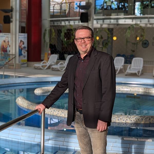Ein Mann mit dunklem Sakko und Hemd steht vor dem gefüllten Erlebnisbecken im Schwimmbad De Bütt.