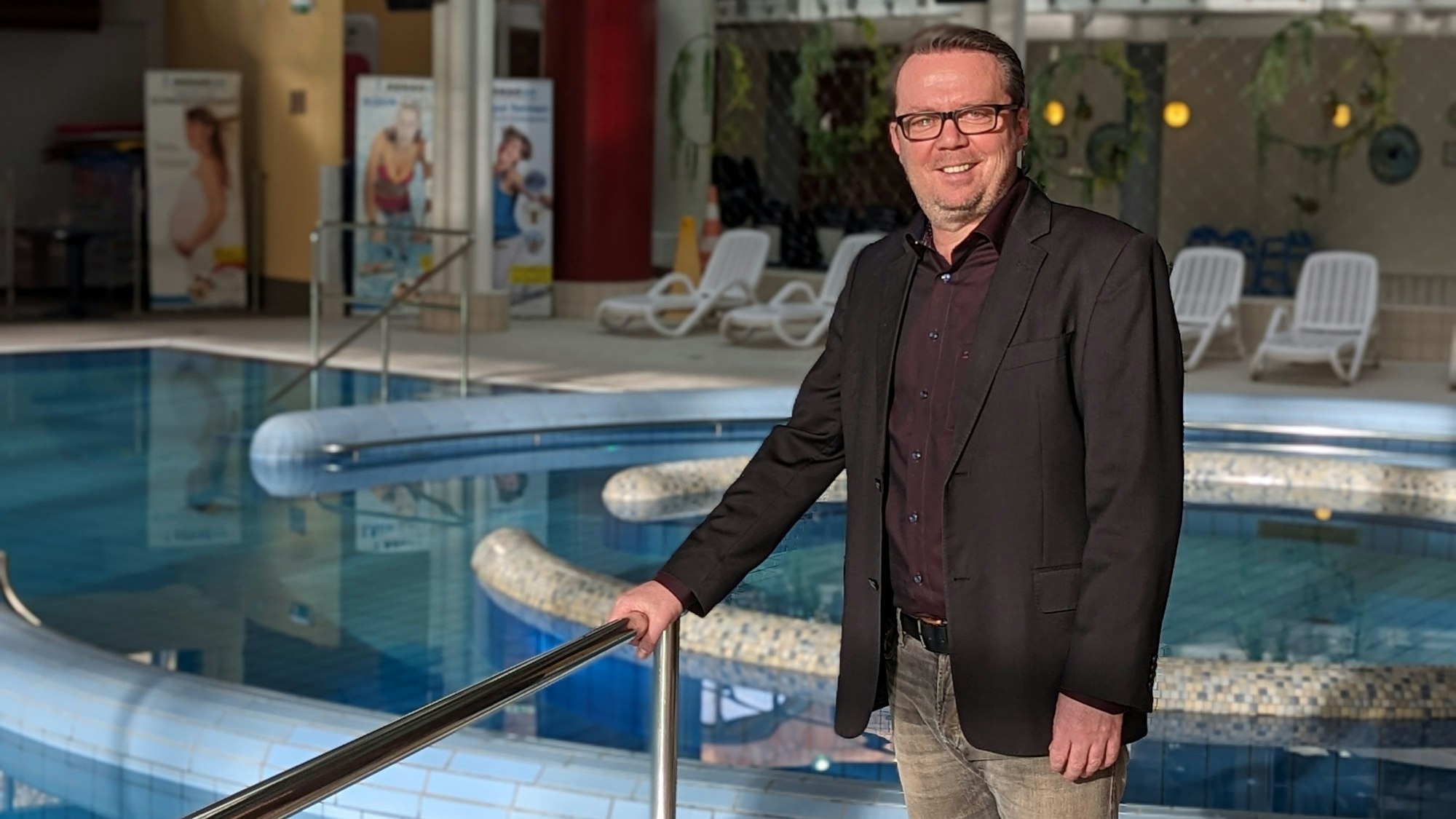 Ein Mann mit dunklem Sakko und Hemd steht vor dem gefüllten Erlebnisbecken im Schwimmbad De Bütt.